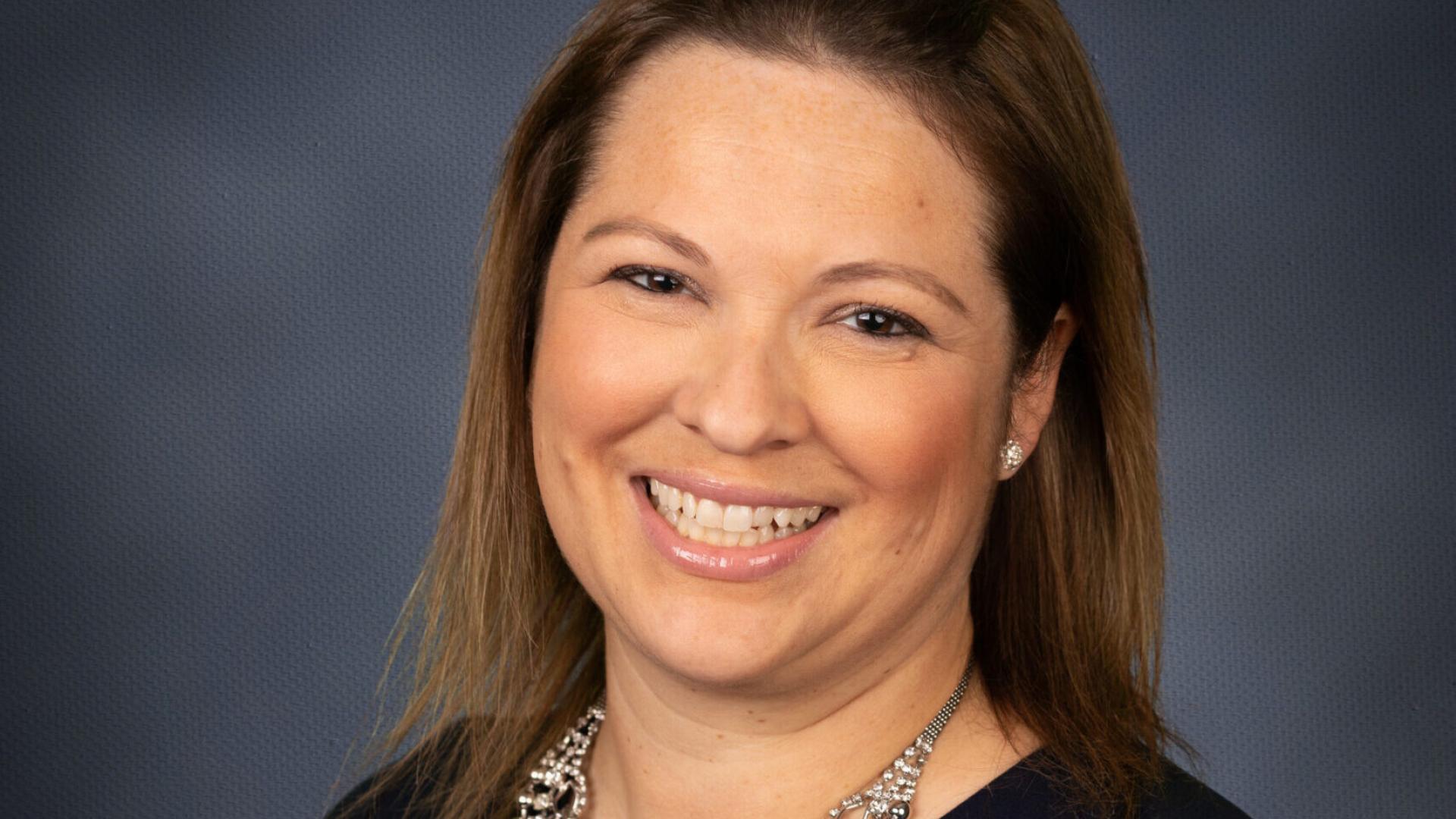 Portrait of a person smiling, wearing a necklace and dark shirt against a grey background.