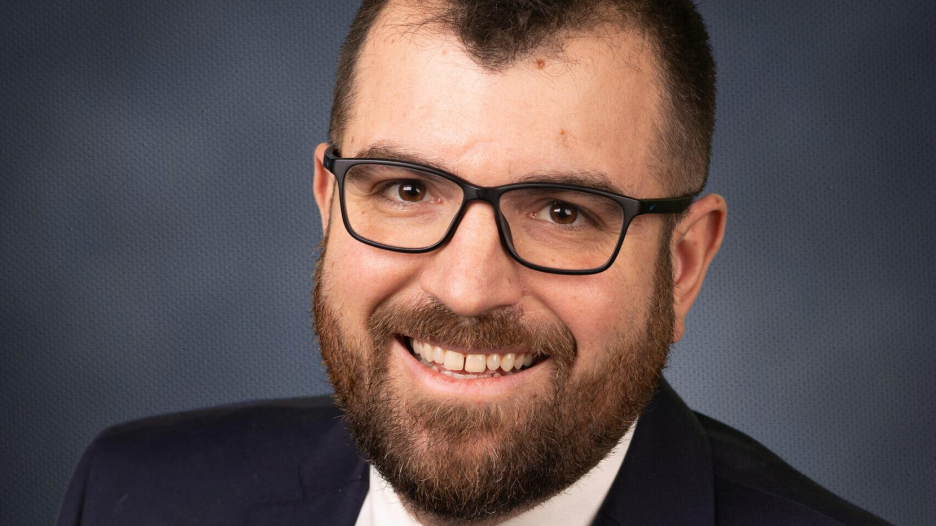 Smiling person in a suit with glasses and a beard, against a dark background.