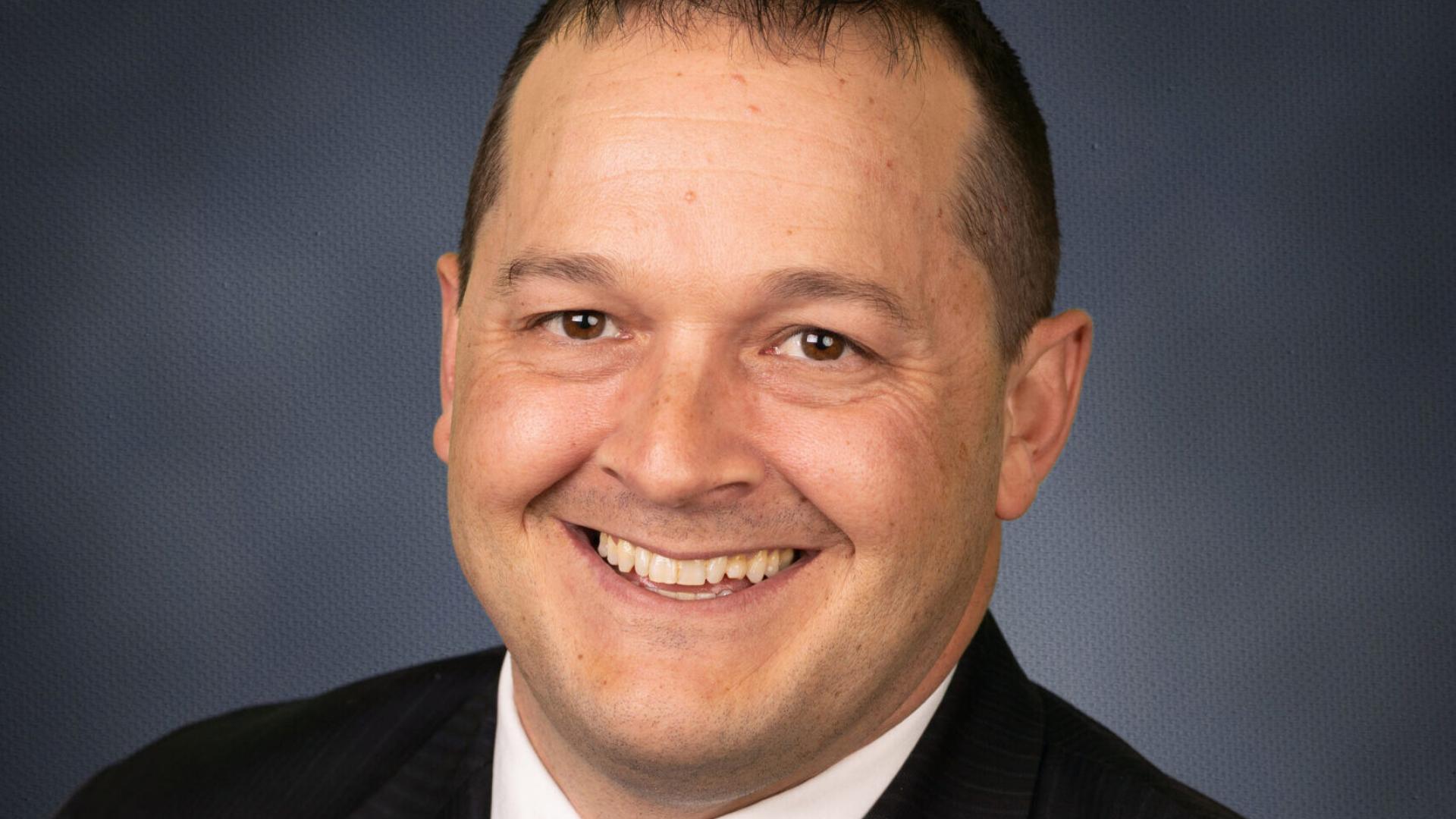 A person smiling, wearing a black suit and blue tie, with a dark blue background.