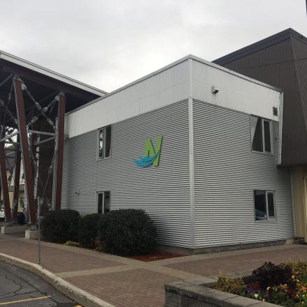 Modern building with a slanted roof, silver siding, and a green-blue logo. Overcast sky.