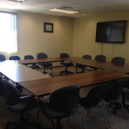 Empty conference room with U-shaped table, chairs, and a wall-mounted TV.