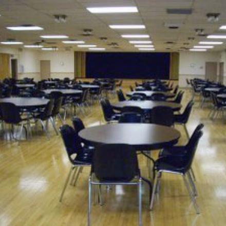 Empty dining hall with round tables and chairs, wooden floor, bright lighting.