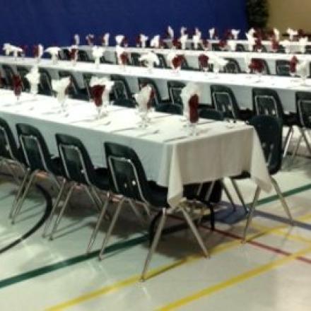 Rows of tables with white tablecloths, set with folded napkins, in a large hall.